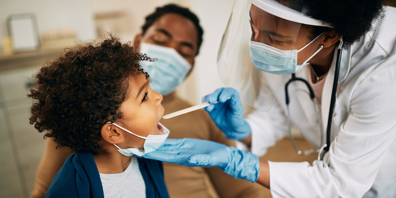 Family doctor examining throat of a small black boy while visiting him at home during coronavirus pandemic.